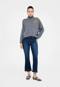 Maglione grigio in maglia con collo alto, caratterizzato da un dettaglio floreale giallo. Abbinato a jeans a zampa di elefante blu scuro e scarpe piatte grigie.