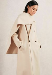 Beige wollen jas met een cape-achtige kraag, dubbele knopen aan de voorkant en zijzakken, met een gladde textuur en een gestructureerd ontwerp.