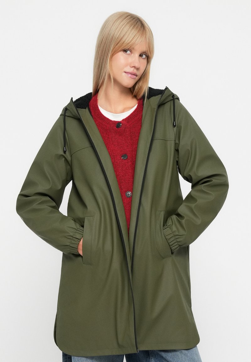 Olijfgroene, middellange regenjas met een capuchon, zwarte voering, voorkant met rits, elastische manchetten en twee zijzakken. Gecombineerd met een rode trui.