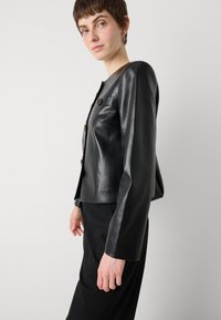 Black leather cropped jacket with a fitted silhouette, featuring long sleeves and two front button details. Smooth texture, minimalistic design.