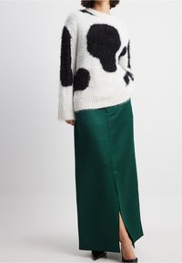 Osoba ubrana w białą, futrzastą sweter z dużymi czarnymi plamami, długą, ciemnozieloną satynową spódnicę z rozcięciem z przodu oraz czarne szpilki.