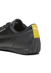Chaussures synthétiques noires avec semelle en caoutchouc texturée, comportant un accent jaune au talon, des perforations et un logo blanc détaillé sur le côté.