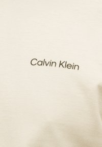 Ύφασμα χρώματος κρέμα με την ένδειξη "Calvin Klein" σε μικρή, κεντραρισμένη, σκούρα γκρι γραμματοσειρά.