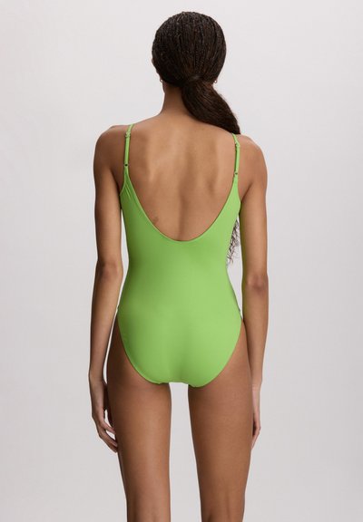 Γυναίκα με πλεγμένα μαλλιά σε χαμηλή αλογοουρά, φοράει ένα ενιαίο μαγιό φωτεινού πράσινου χρώματος με ρυθμιζόμενους ιμάντες, από πίσω.