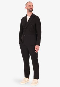 Blazer nero doppiopetto con rever, abbinato a pantaloni coordinati e sneakers color crema. Tessuto morbido, vestibilità su misura, design minimalista.