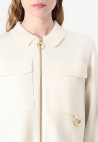 Κρέμα σακάκι με κλασικό γιακά, χρυσό φερμουάρ, δύο τσέπες στο στήθος και διακοσμητικό χρυσό δαχτυλίδι στο πλάι. Απαλή υφή.