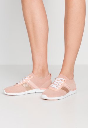 Stopy noszące jasnoróżowe dzianinowe buty sportowe z białymi podeszwami i sznurówkami, z błyszczącym różowo-złotym paskiem po boku, stojące na białej podłodze.