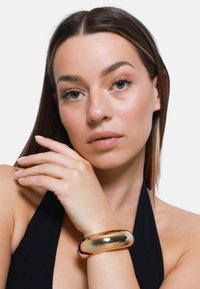 Armband aus goldenem Metall, mit glattem und geschwungenem Design, am Handgelenk getragen, mit glänzender Oberfläche und nahtloser Struktur.