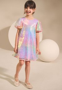 Abito arcobaleno scintillante con paillettes, maniche corte a sbuffo e orlo arrotondato. Abbinato a scarpe piatte scintillanti. Sfondo a tinta unita.
