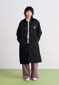 Personne se tient avec les mains dans les poches, portant un long manteau noir, une chemise blanche, un pantalon mauve ample et des chaussures blanches devant un fond uni.