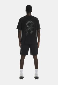 Tricou negru din bumbac cu un design grafic pe spate, asortat cu pantaloni scurți negri. Modelul poartă șosete albe și pantofi sport gri deschis.