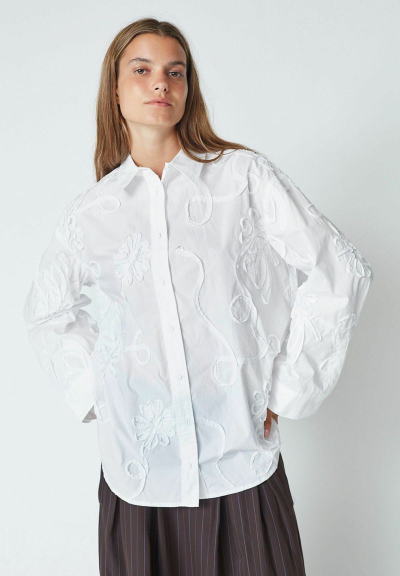 Biele button-up košeľa s kvetinovou výšivkou, oversized rukávy, textúrované detaily a zakrivený lem, v kombinácii s hnedou skladanou sukňou.