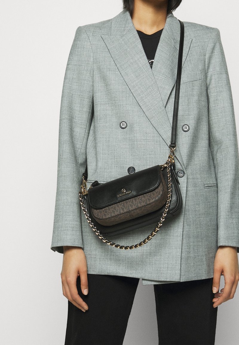 Personne portant un blazer croisé gris clair et un pantalon noir, portant un sac bandoulière noir et marron avec une lanière chaîne dorée.