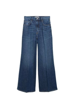 Personne portant un blazer en cuir noir sur un haut noir et un jean bleu taille haute à jambes larges, debout avec une main dans la poche devant un fond uni.