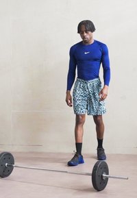 Sportininkas, vilkintis mėlynos spalvos ilgomis rankovėmis Nike marškiniais ir leopardų rašto šortais, stovi šalia juodo štangos ant šviesaus grindų paviršiaus.