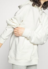 Sudadera con capucha color crema, con bolsillo delantero, capucha con cordón y puños acanalados. Tela suave, ajuste relajado y detalles de costuras visibles.