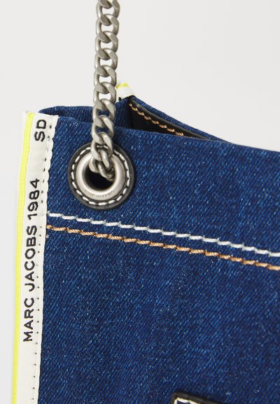 Geanta de denim cu un umăr din lanț, având accente galbene, cusături albe și o etichetă cu logo. Material texturat albastru cu elemente metalice argintii.