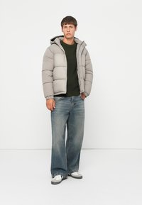 Puffer-jakke i lys grå, med glidelås og knappelukking, festet hette og quiltet design. Matchende med vide blå jeans og sneakers.
