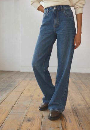 Blaue, weit ausgestellte Jeans aus Denim mit hoher Taille, die ein dezentes gesprenkeltes Muster und ein traditionelles Fünf-Taschen-Design aufweisen, getragen mit braunen Schuhen.