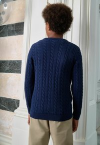 Tumesinine kootud kampsun palmikumustriga, ümar kaelus ja soonikkäärid, koos heledate pükstega.