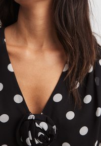 Blusa de tela negra con lunares blancos, con un escote en V profundo y un acento de rosa de tela en el pecho. Textura suave.