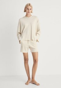 Jasno beżowa bluza z kapturem i dopasowane szorty, oba wykonane z miękkiego materiału. Bluza ma szerokie rękawy, a szorty mają elastyczną talię z sznurkiem.
