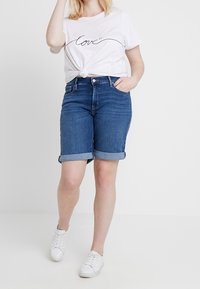 Denimshortsit, joissa käännetyt lahkeet, keskisininen pesu, yhdistetty valkoiseen puuvillapaitaan, jossa on kursiivilla kirjoitettu "love"-kuvio. Valkoiset tennarit.