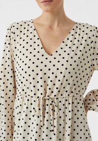 Sukienka w kremowym kolorze w czarne kropki, z dekoltem w serek, marszczoną talią z troczkiem i długimi rękawami. Gładka faktura tkaniny.