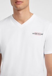 Valge puuvillane V-kaelusega T-särk, millel on vasakul üleval väike punane ja must "Guess Jeans" logo; sile tekstuur ja lõdva istuvusega.