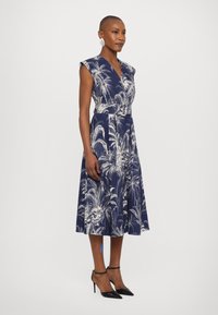 Rochie bleumarin cu imprimeu floral, decolteu în V, mâneci scurte, talie ajustată și fustă evazată, asortată cu tocuri negre stiletto.