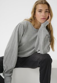 Pull gris avec des manches bouffantes texturées ; corps lisse ; encolure arrondie ; associé à un jean foncé. Mannequin assis, le menton reposant sur la main.