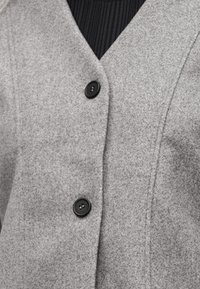 Κοντινό πλάνο ενός γκρι μάλλινου παλτού με δύο μαύρα κουμπιά πάνω από μια μαύρη ριγέ μπλούζα.