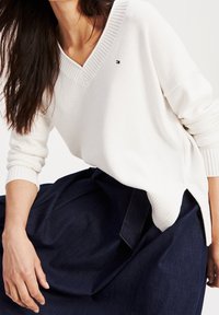 Pull en maille blanche avec un col en V, des poignets côtelés et des fentes sur les côtés, associé à une jupe en denim foncé. Petit logo brodé sur la poitrine.