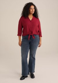 Blusa rossa con texture, con nodo frontale e bottoni, abbinata a jeans svasati blu scuro e calzature nere, posta su uno sfondo neutro.