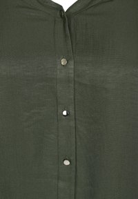 Chemise vert olive avec un léger éclat, présentant un devant à boutons et des boutons métalliques pour la fermeture. Texture de tissu lisse.