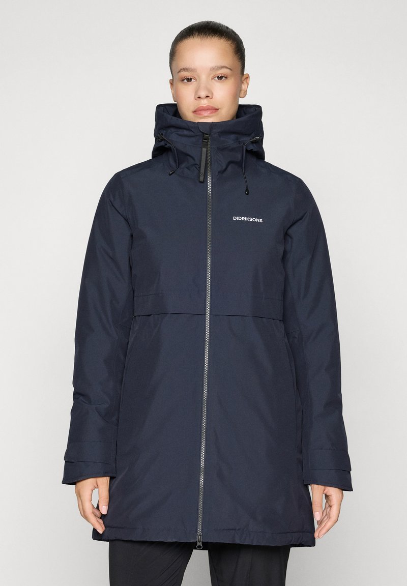 Αδιάβροχο παρκά σε ναυτικό μπλε χρώμα με ψηλό γιακά, ρυθμιζόμενο καπό, φερμουάρ μπροστά ολόκληρο και πλαϊνές τσέπες. Ομαλή υφή, μεσαίου μήκους σχεδίαση, λογότυπο στο στήθος.