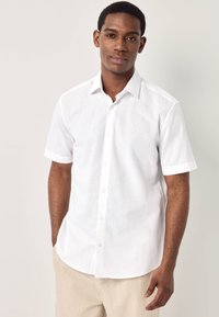 Giovane uomo con una camicia bianca a maniche corte abbottonata e pantaloni beige, in piedi con una mano in tasca davanti a uno sfondo semplice.