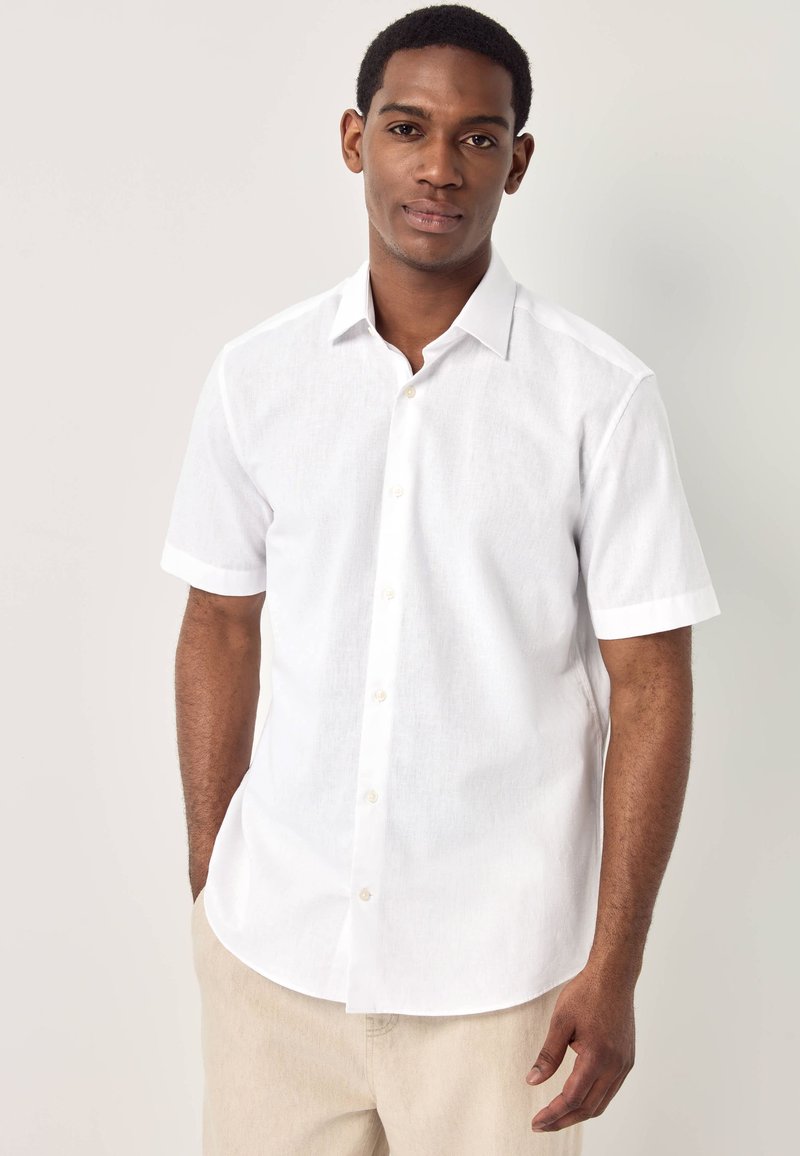 Giovane uomo con una camicia bianca a maniche corte abbottonata e pantaloni beige, in piedi con una mano in tasca davanti a uno sfondo semplice.