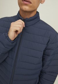 Giacca imbottita blu navy con colletto alto, chiusura con zip e motivi di cucitura orizzontali. Il materiale sembra liscio con una texture leggermente imbottita.