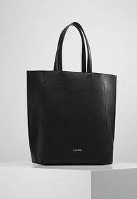 Czarna skórzana torba typu tote z dwoma uchwytami, przestrzenny prostokątny kształt, minimalne przeszycia oraz małe metalowe logo na dole.
