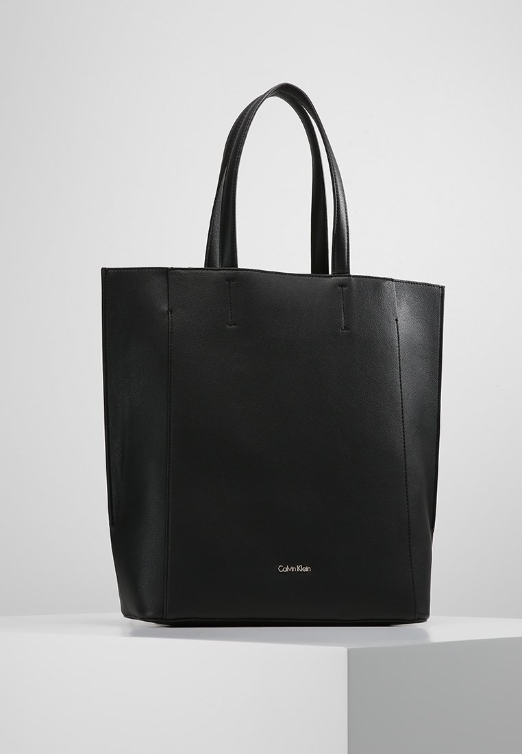 Czarna skórzana torba typu tote z dwoma uchwytami, przestrzenny prostokątny kształt, minimalne przeszycia oraz małe metalowe logo na dole.