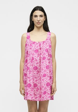 Γυναίκα με μακριά σκούρα μαλλιά, που φοράει ένα αμάνικο ρόζ και λευκό φόρεμα με λουλουδάτο μοτίβο μέχρι το γόνατο, στέκεται μπροστά σε απλό φωτεινό φόντο.