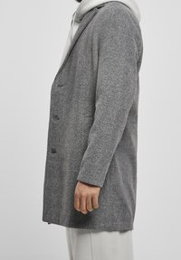 Grå herringbone-frakke med en struktureret design, knaplukning og klapkrave, lagd over en lys hoodie, der fremhæver et tekstureret stof.