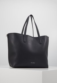 Czarna skórzana torebka typu tote z podwójnymi ramiączkami, lekko zaokrąglonym górnym brzegiem i małym logo "Karl Lagerfeld" z przodu, na białym postumencie.