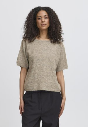 Kvinde med krøllet hår iført en kortærmet beige sweater og løstsiddende sorte bukser, stående mod en ensfarvet lys baggrund.