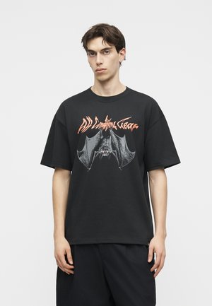 Νέος άνδρας που φοράει μαύρο φαρδύ T-shirt με μεγάλο σχέδιο νυχτερίδας και πορτοκαλί κείμενο στο στήθος, στέκεται μπροστά σε ένα απλό λευκό φόντο.