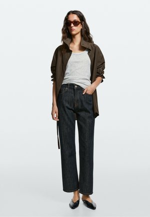 Frau trägt übergroße dunkelbraune Jacke, graues T-Shirt, dunkle weit geschnittene Jeans, schwarze spitze Ballerinas und große braune Sonnenbrille, stehend.
