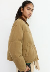 Ung kvinde med mørkt hår sat op i en hestehale iført en brun polstret jakke mod en ensartet hvid baggrund, der ser på kameraet.