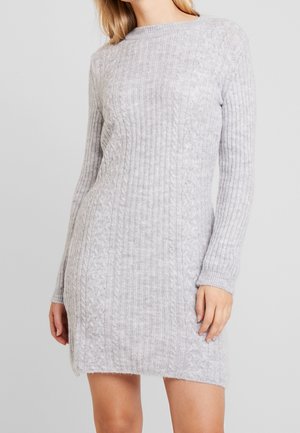 Kvinde iført en lysegrå, langærmet, knælængde strikket sweaterkjole med kabel- og ribmønstre mod en ensfarvet baggrund.