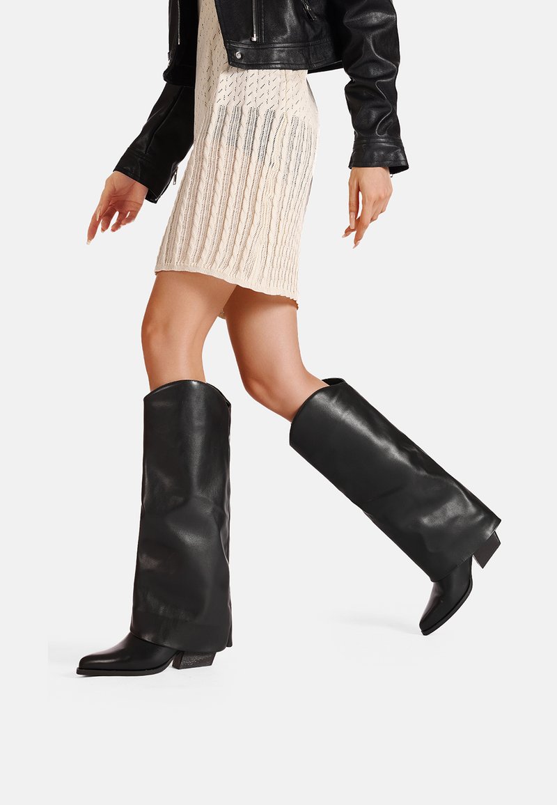 Stivali neri al ginocchio in pelle lucida, con un design elegante, punta affusolata e tacco a blocco. Indossati con un vestito in maglia color crema.